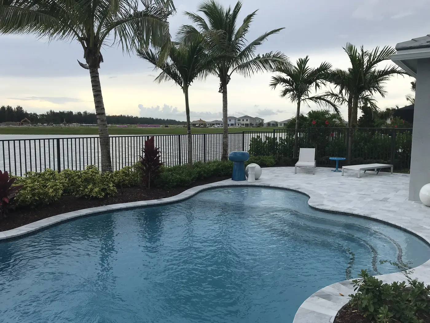 A curving backyard pool with young palms, a white stone patio and lounge chairs at a Minto model home in Westlake, with a lake and new homes in the distance