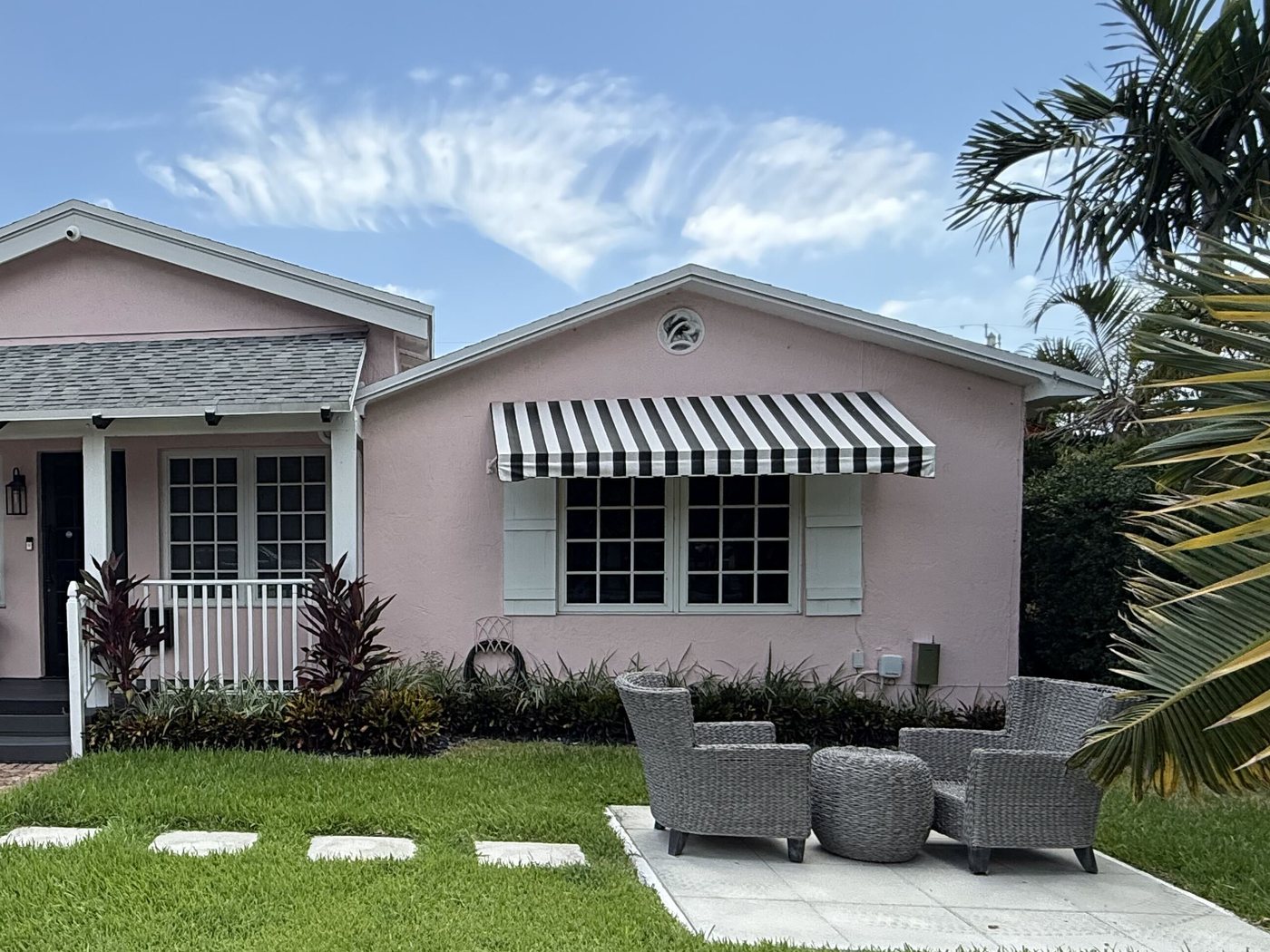 Renovated pastel beach cottage with a black-and-white striped awning and a wicker seating area