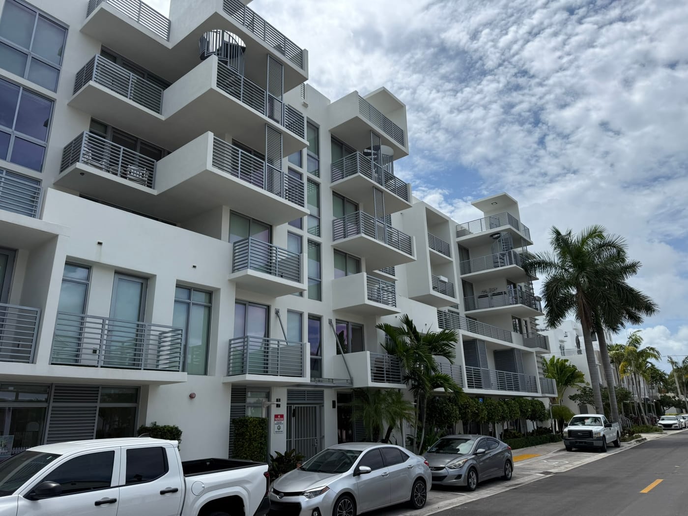 A mid-rise white condominium building with stacked balconies along a street lined with parked cars