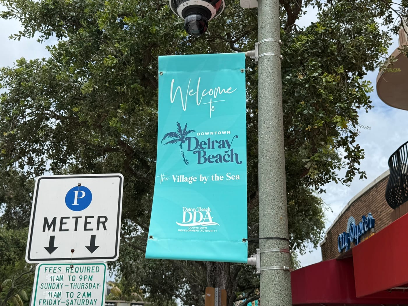 A downtown parking meter sign with posted fee hours beside a Downtown Delray Beach banner