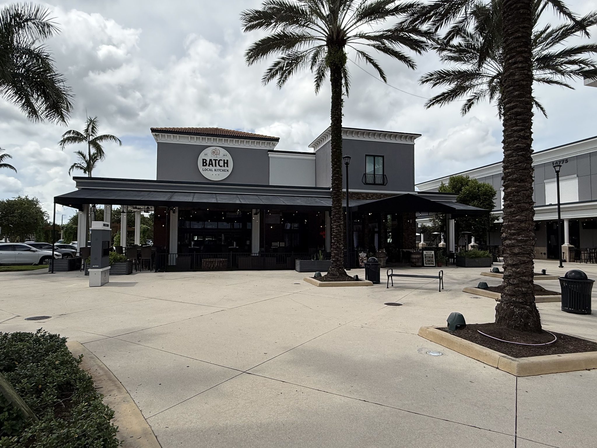 Batch Local Kitchen at Delray Marketplace, a two-story building with a black awning over a covered patio, framed by palms