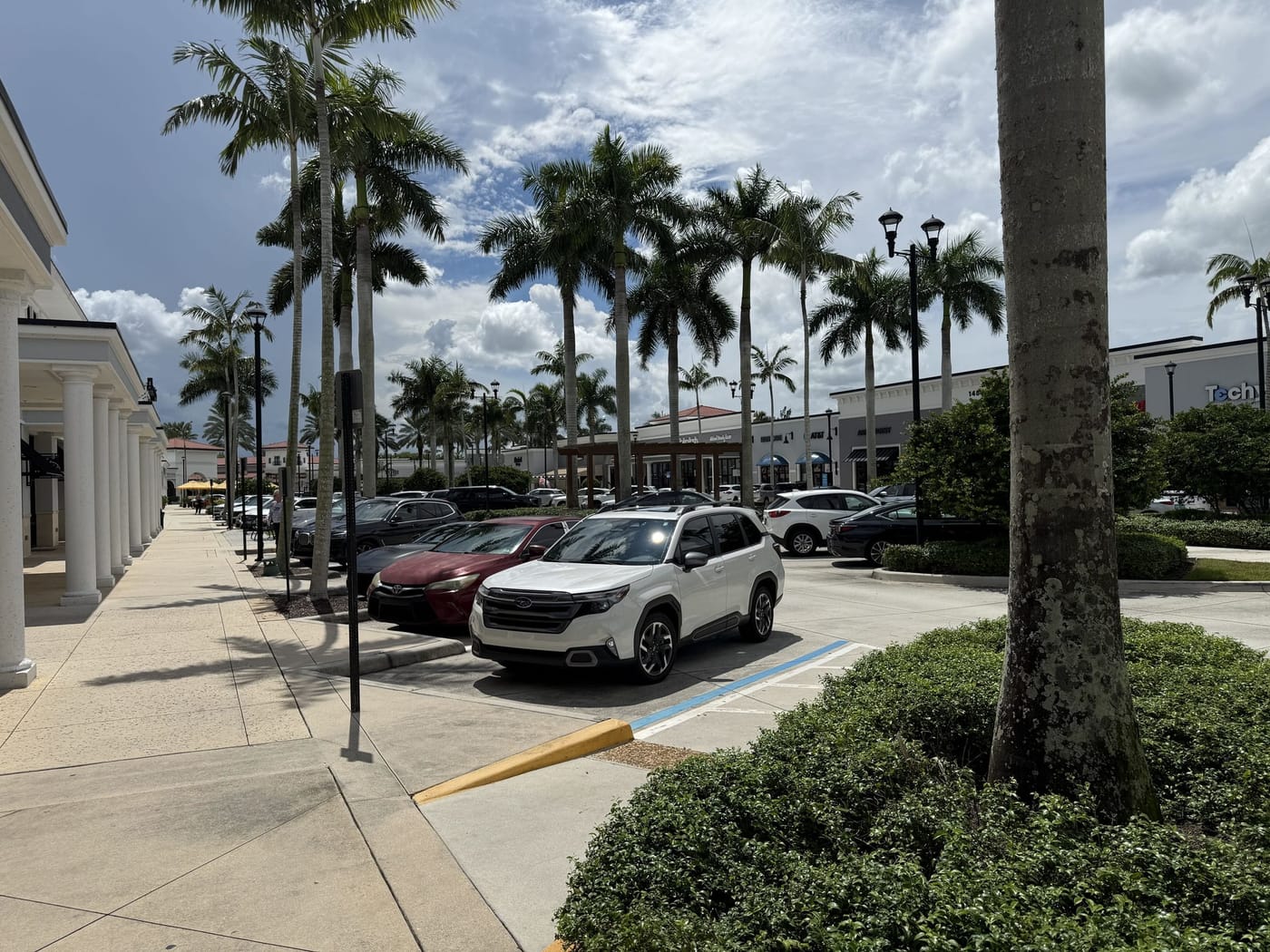 A covered colonnade sidewalk along storefronts with the surface parking field beside it