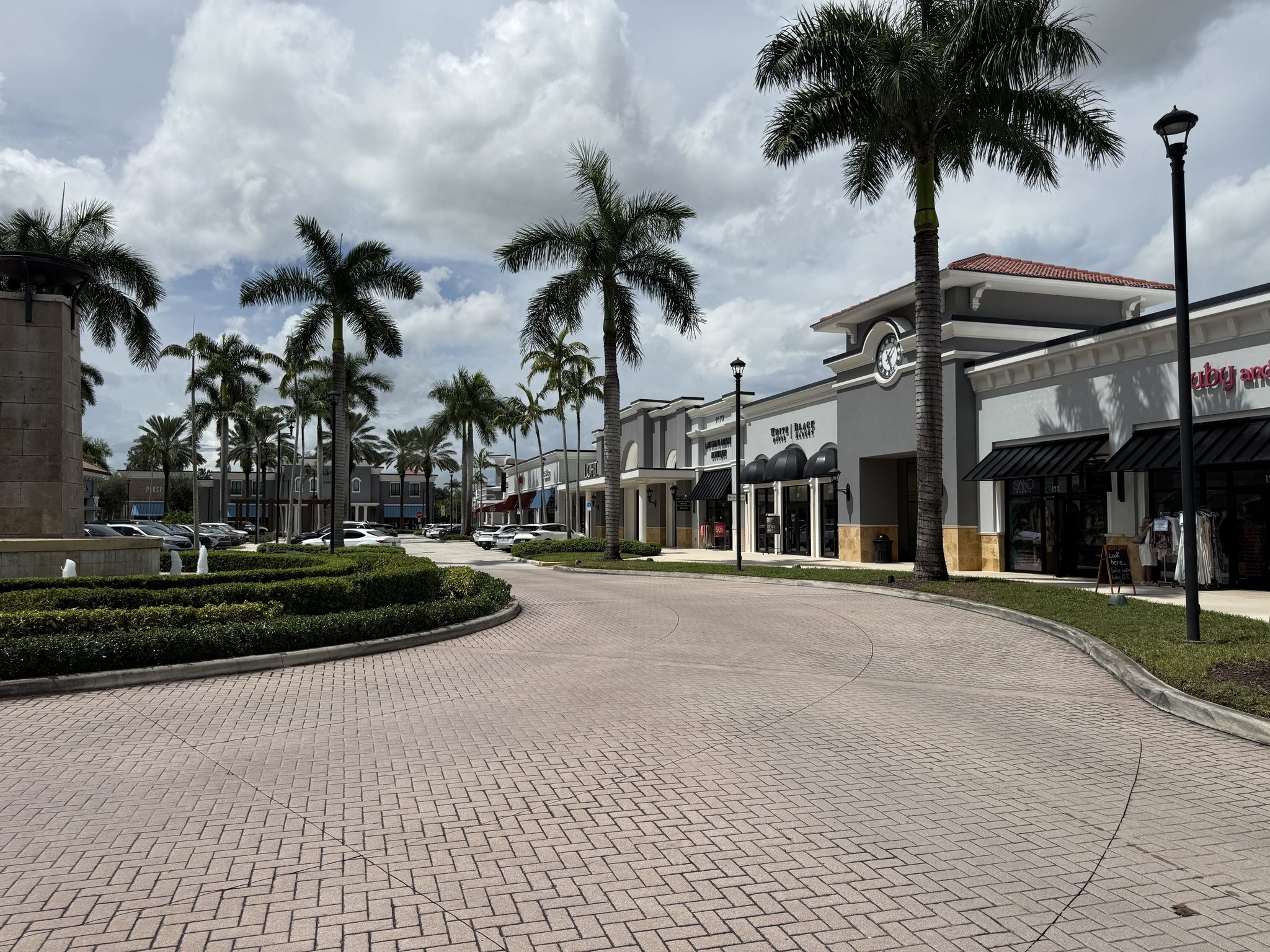 Storefronts along a curving brick drive at Delray Marketplace, with royal palms and lamp posts